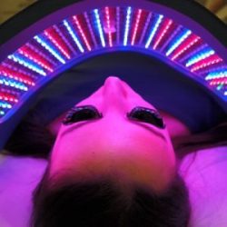 A person holding a red light therapy device to their face, with a peaceful expression as the device emits red light which is known to help with various skin conditions and promote overall skin health